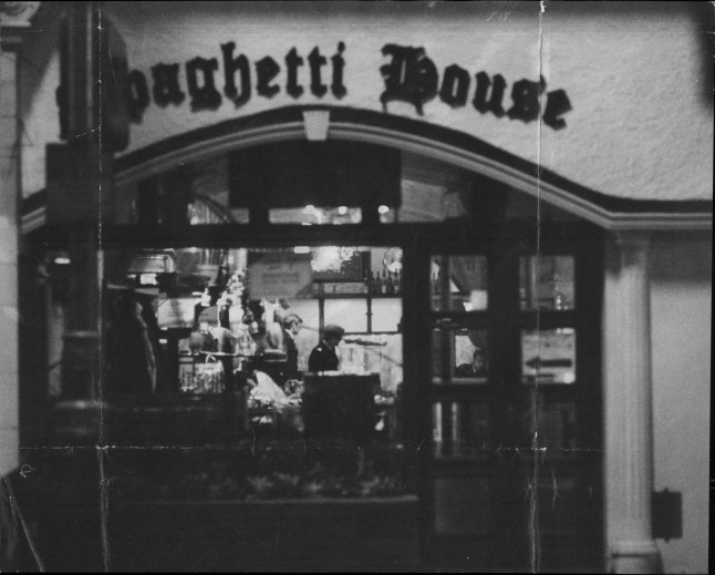 A photo of a print photo in black and white of 'Spaghetti House' (text shows this name above the glass front of the restaurant). The photo is from the outside, so the glass door and front can be seen, revealing the inside of the restaurant which is lit up. The Met Police can be seen inside, and this was taken at the time when the hostages were being held downstairs in the basement.